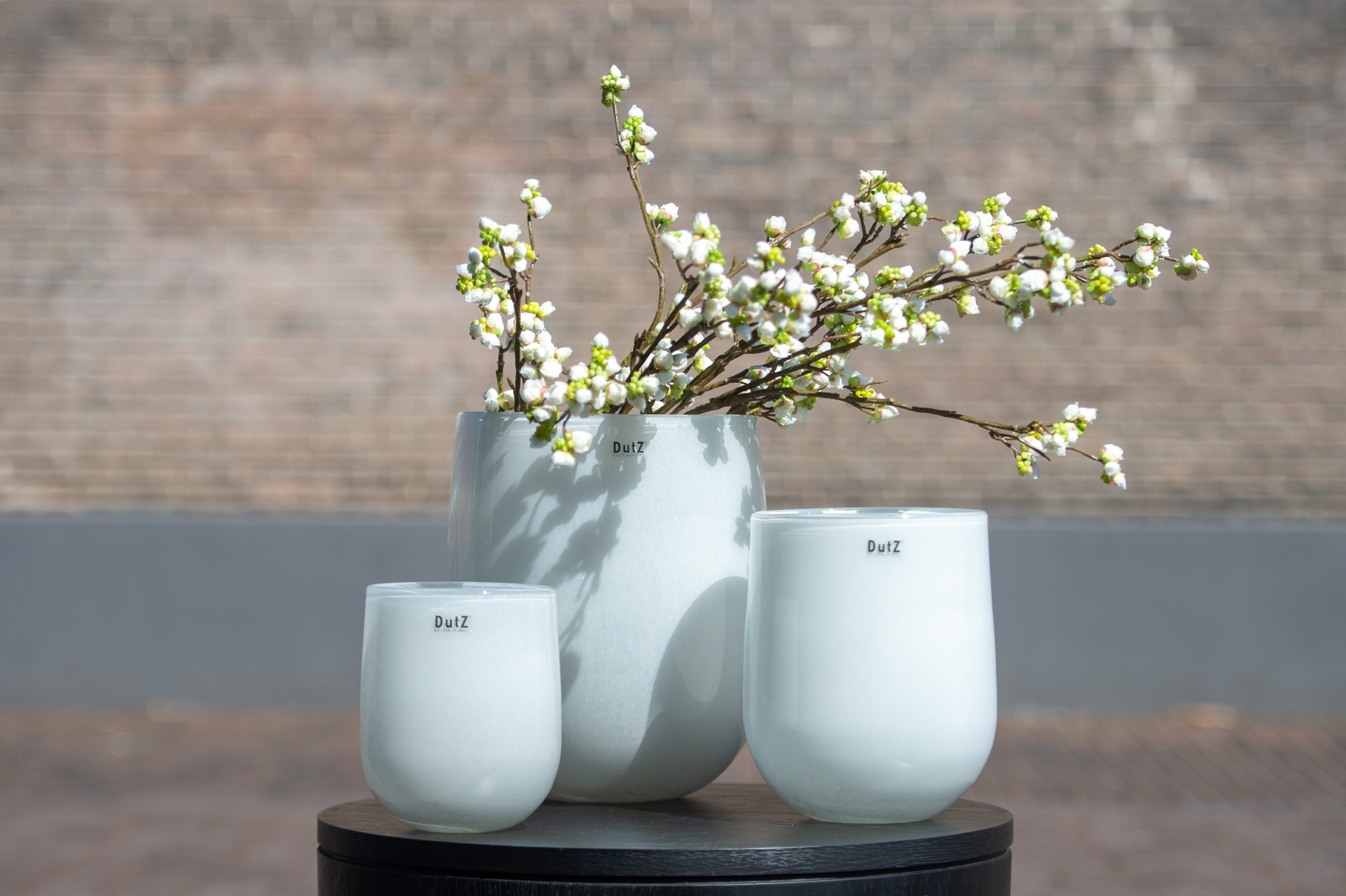 Three DutZ Collection white barrel vases. In the biggest one is a blossom branch. It stands on a round tabel