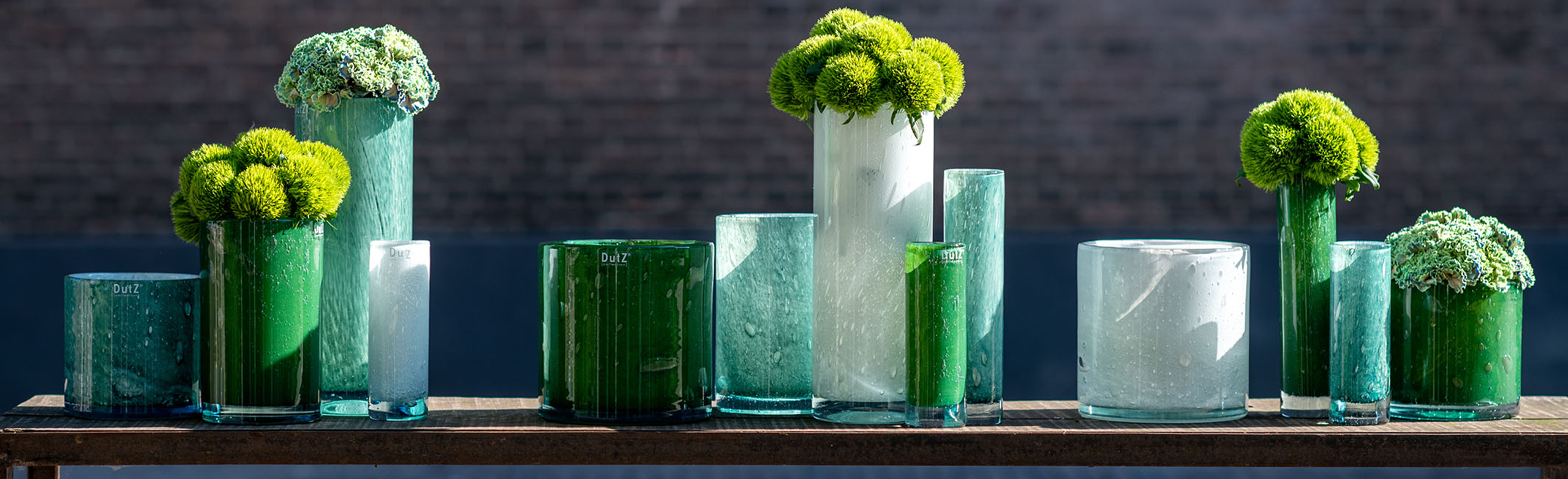 DutZ recycled glass cylinders in the colour green and wihite. On a small table are cylinders in different sizes. Some are filled with flowers